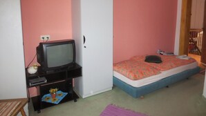 Quarto com cama, guarda-roupa, televisão sobre um suporte e uma pequena mesa com um vaso e flores.