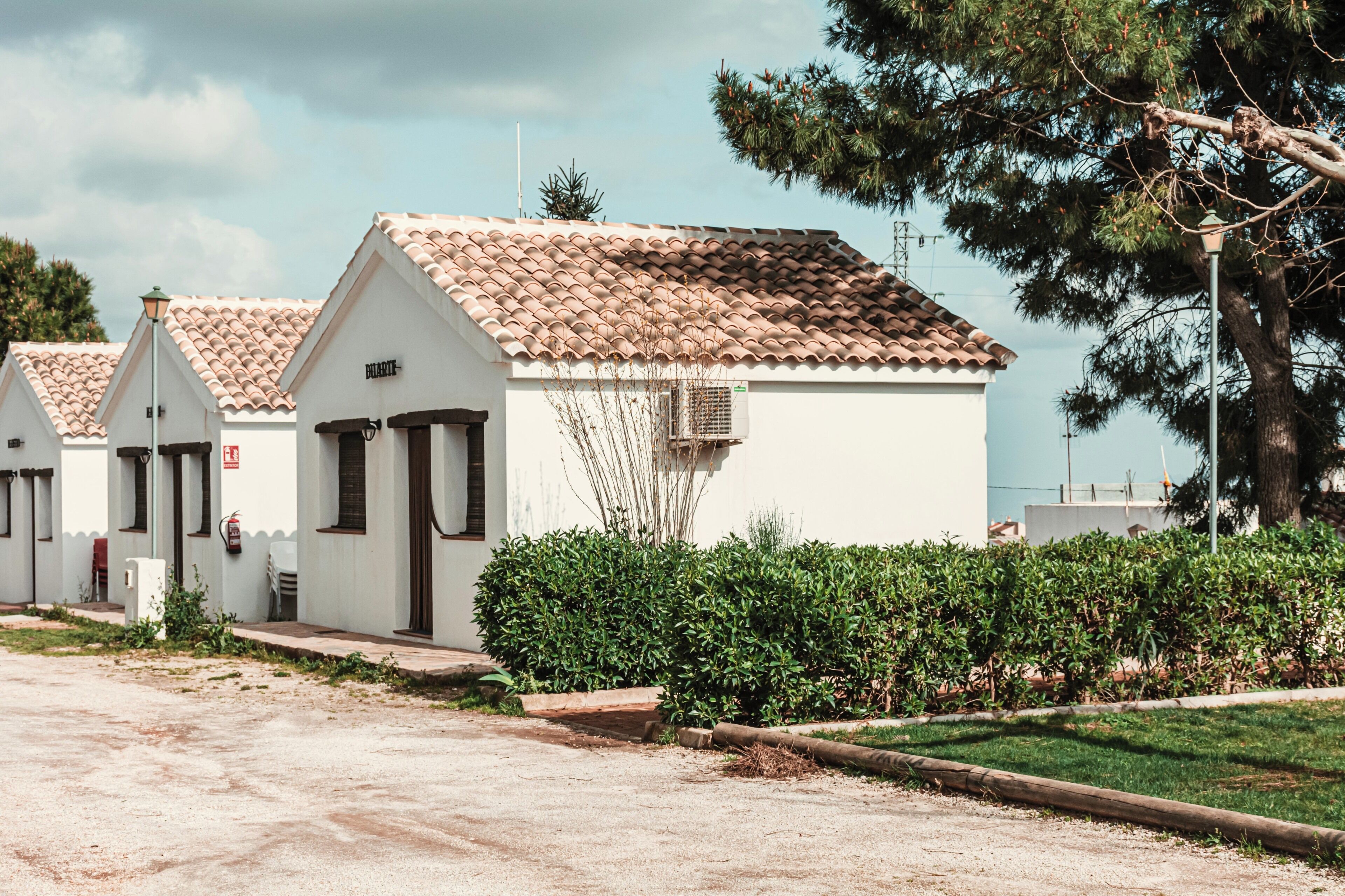 A row of white buildings with brown tiled roofs, a paved path, and a hedge in the foreground.