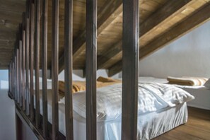 Quarto com teto inclinado e vigas de madeira, apresentando uma cama com roupa de cama branca e um travesseiro.