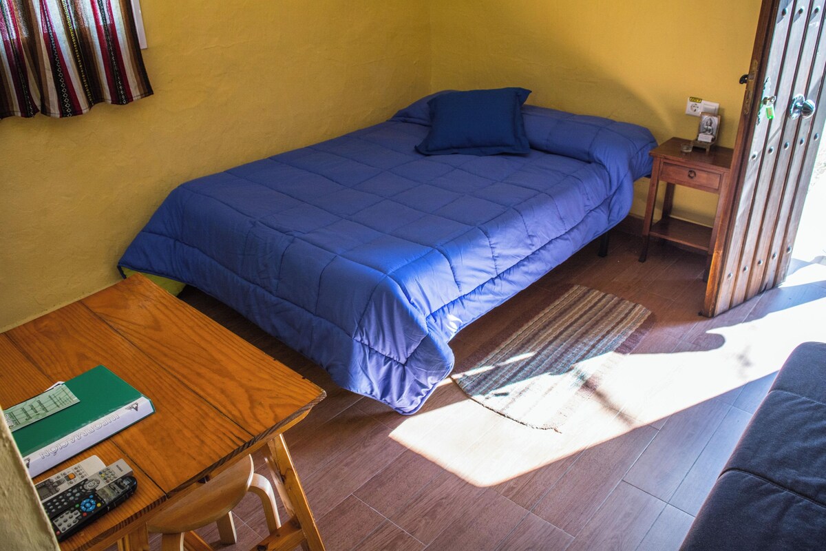 Quarto de casal com uma colcha azul, uma mesa de madeira com um controle remoto e uma pequena mesa de cabeceira com uma luminária.