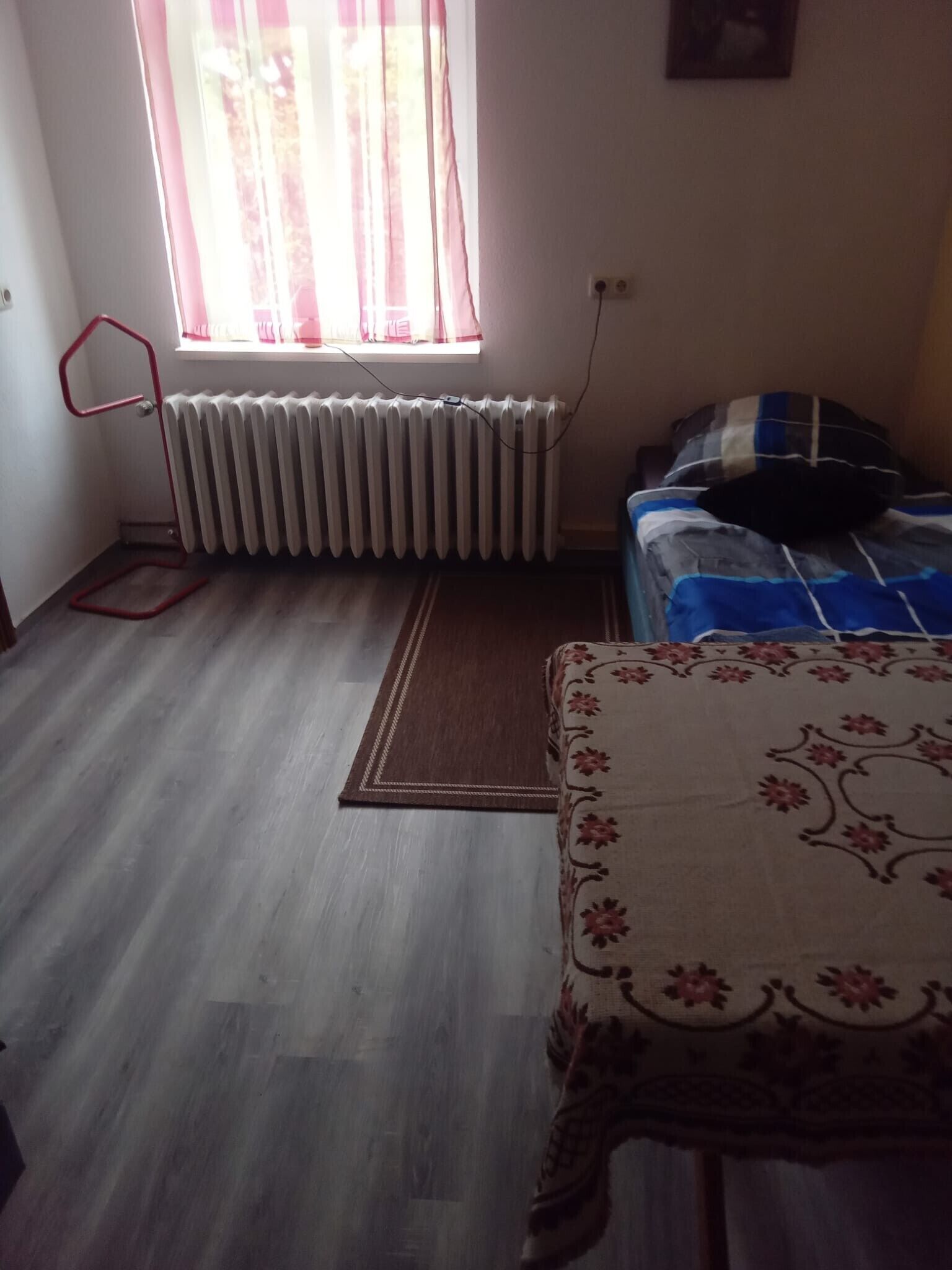 Una habitación con un radiador, una ventana con cortinas, una cama con colcha azul y blanca a rayas, y una alfombra estampada en el suelo.