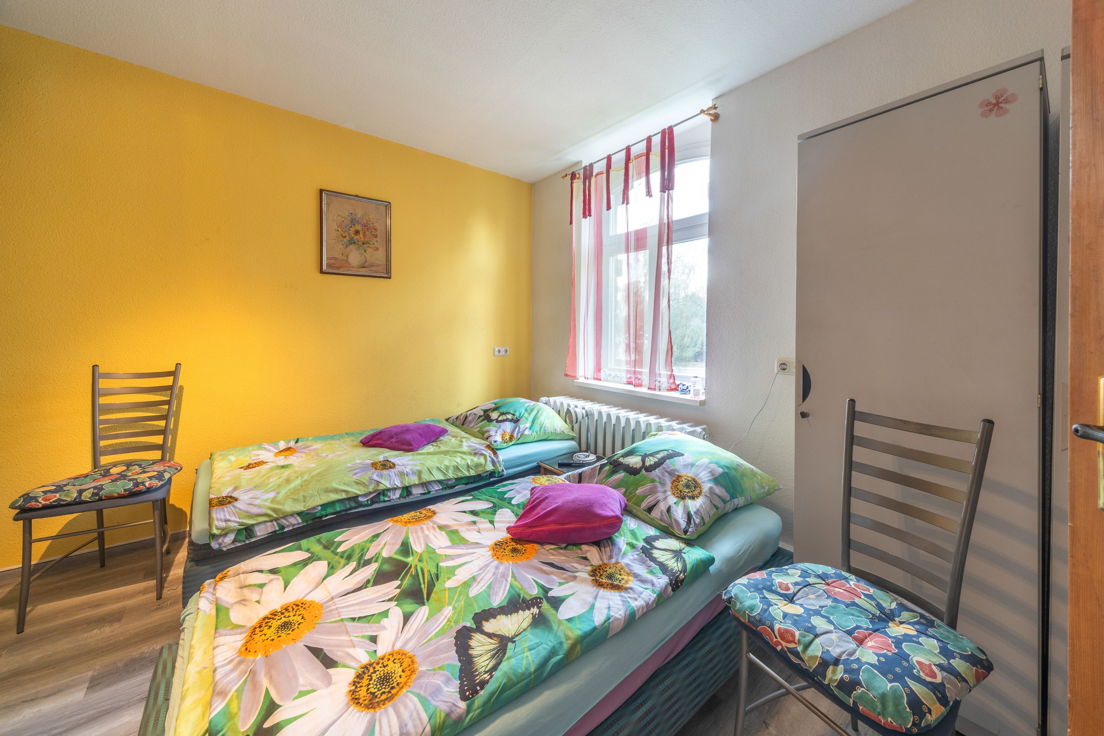 A room with a bed, two chairs, a radiator, a window with curtains, and a door.