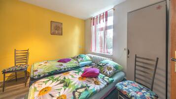 Ein Zimmer mit einem Bett, zwei Stühlen, einem Heizkörper, einem Fenster mit Vorhängen und einer Tür.