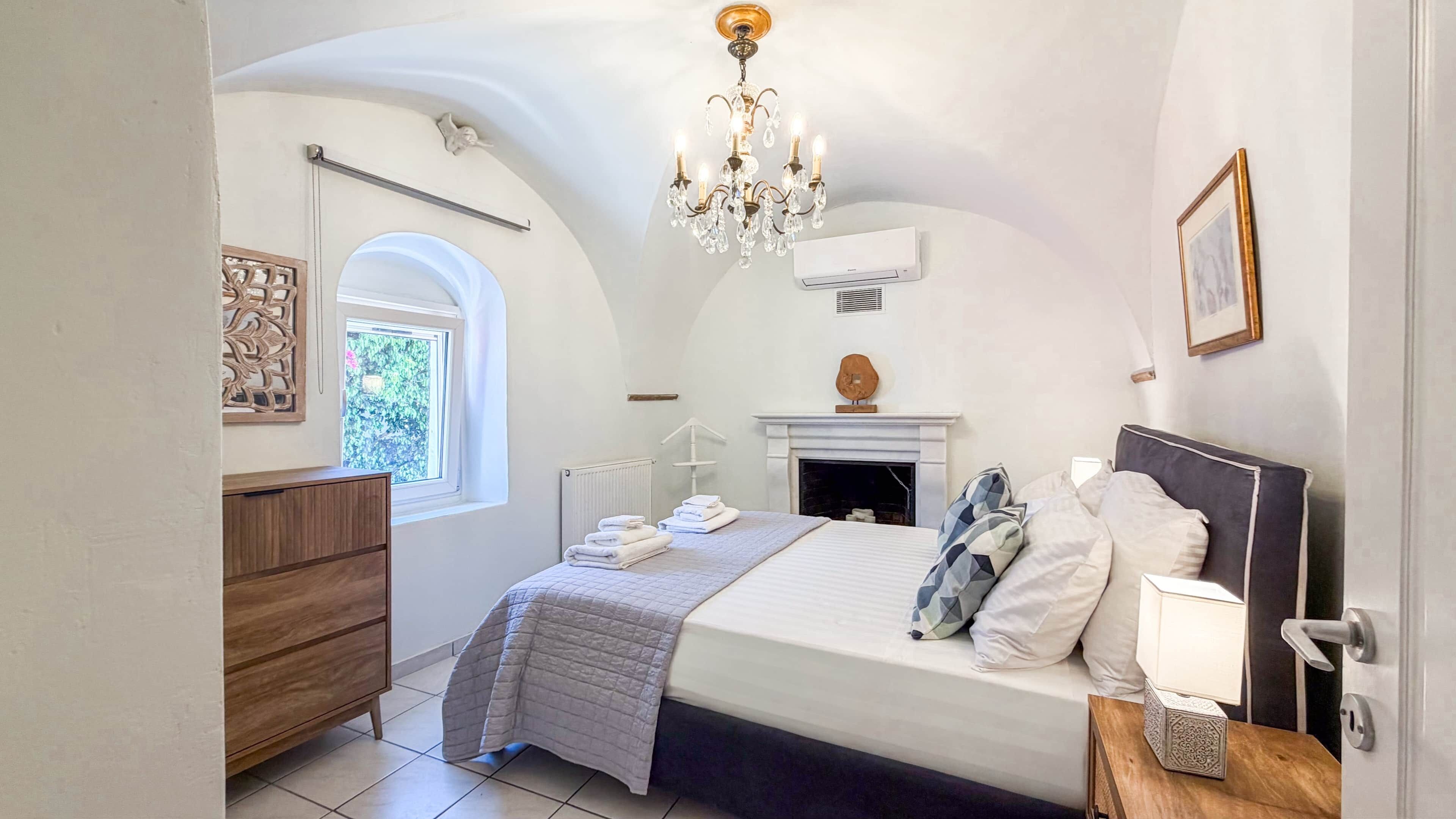 A bedroom with a bed, a dresser, a chandelier, a fireplace, and a window.