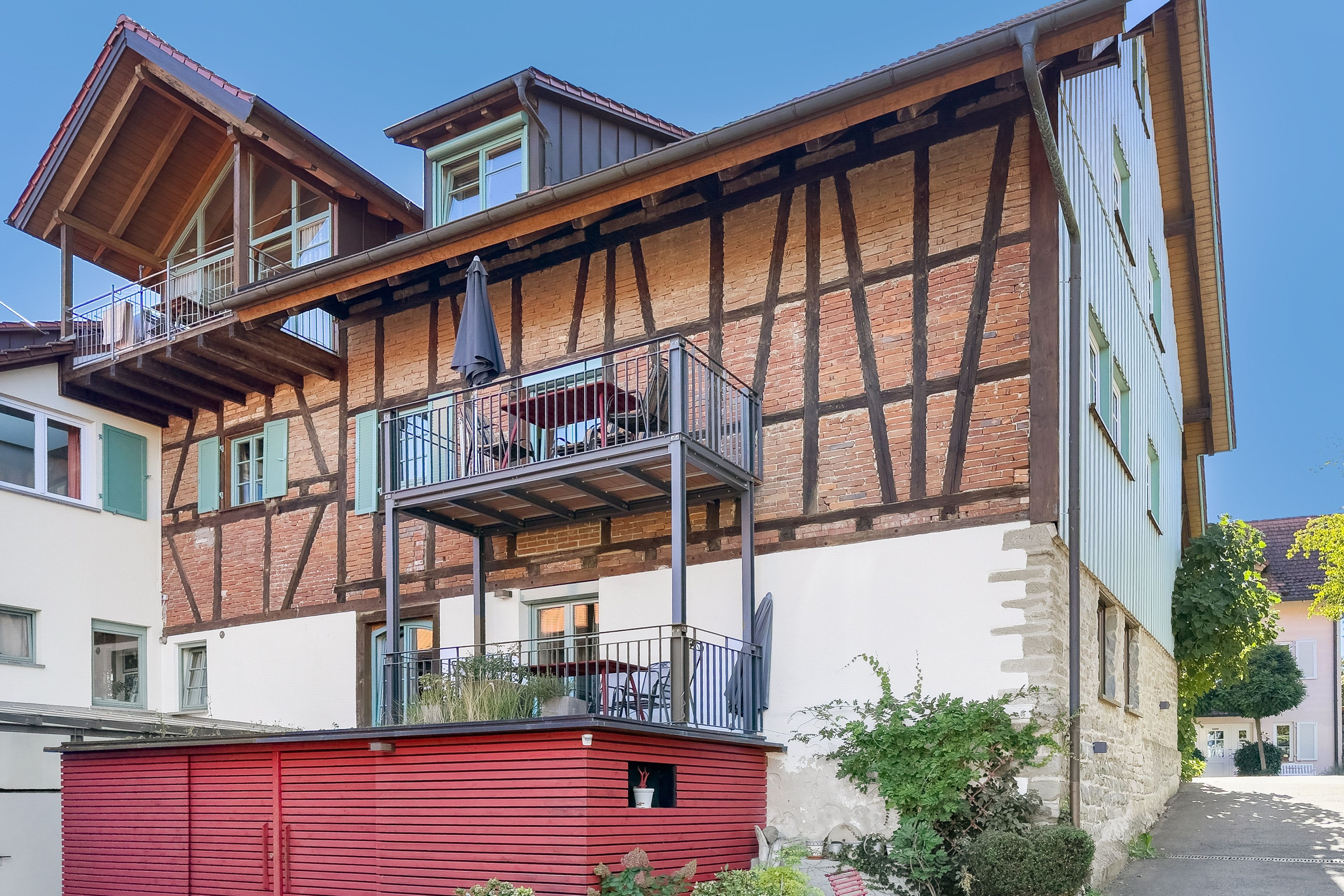 Un immeuble à plusieurs étages avec une charpente en bois apparent, une toiture rouge et un balcon doté d’une rambarde en métal.