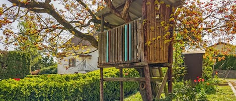 A wooden treehouse structure with a slanted roof, supported by wooden beams, surrounded by lush greenery and flowering trees.