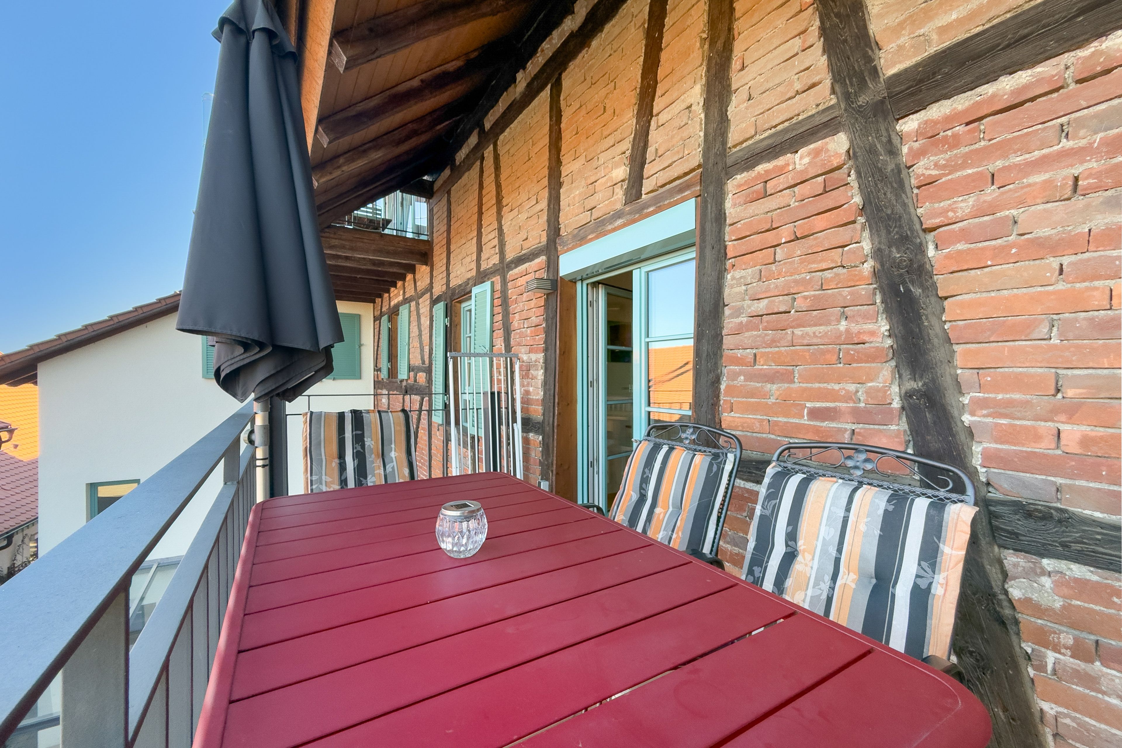 A red table with metal chairs on a balcony with a brick wall and wooden beams.