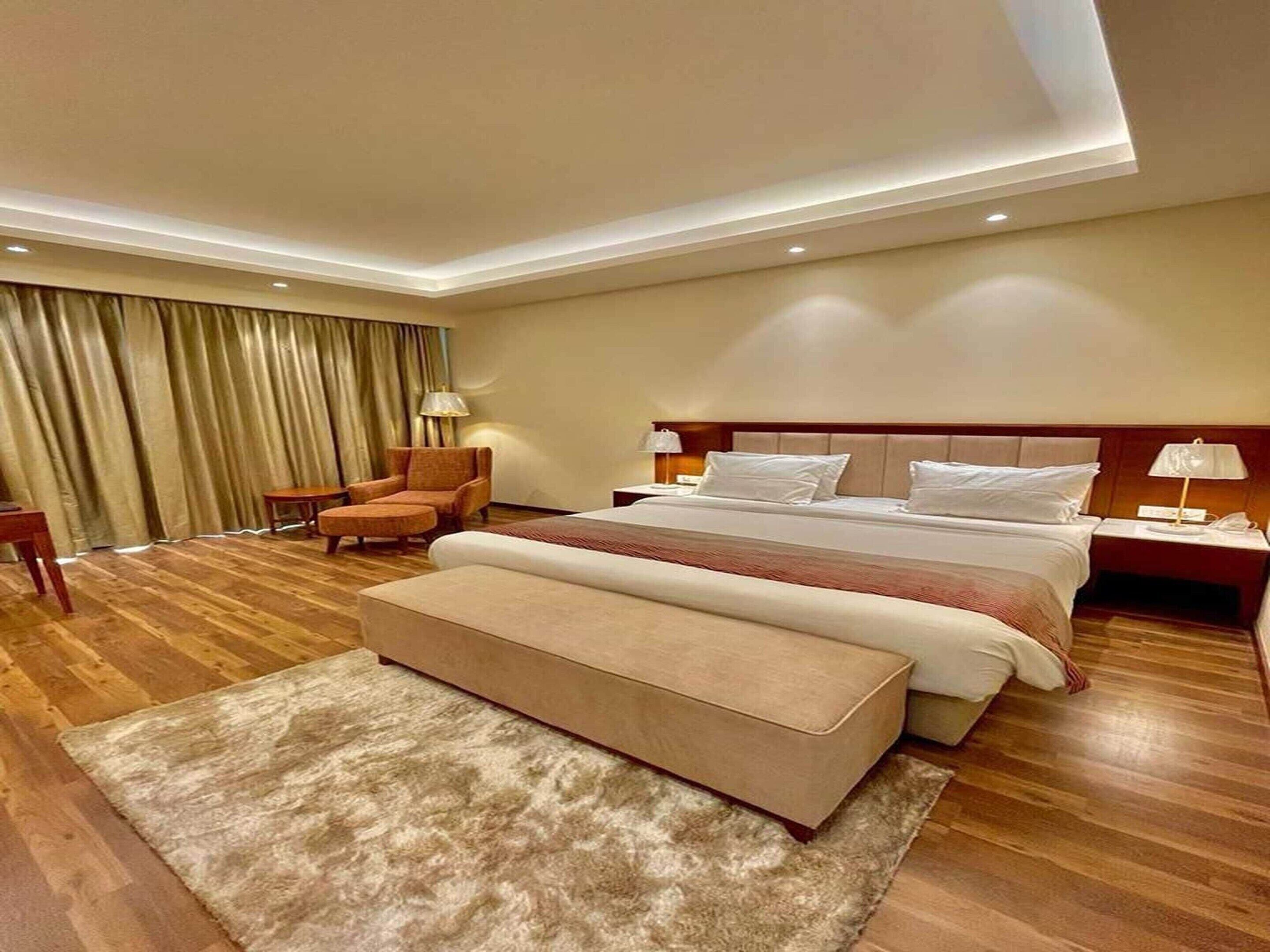 Une chambre d’hôtel dotée d’un grand lit, d’un coin salon avec un fauteuil et un pouf, d’un bureau et d’une fenêtre avec des rideaux.