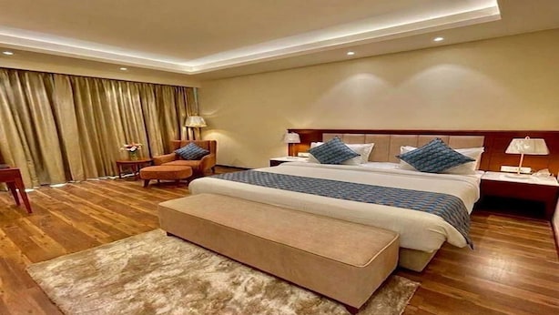 Habitació d'hotel amb un llit gran, zona de seients amb una cadira i otomana, escriptori i finestra ampla amb cortines.