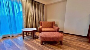 Una habitació amb sòl de fusta, un butac armchair vermell, una taula rodona petita i un gran finestral amb cortines.