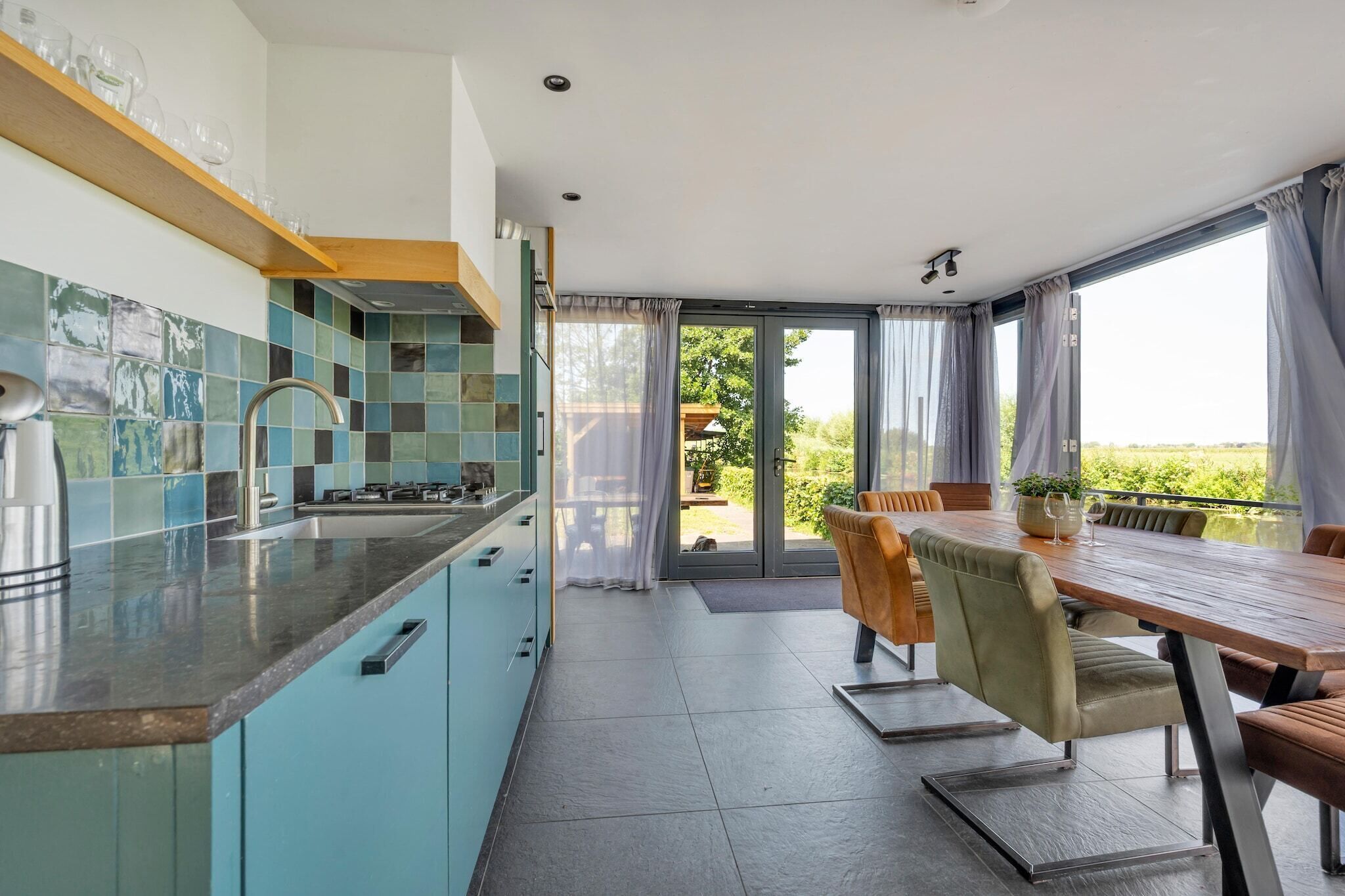 A modern kitchen with a dining area, large windows, and a view of greenery.