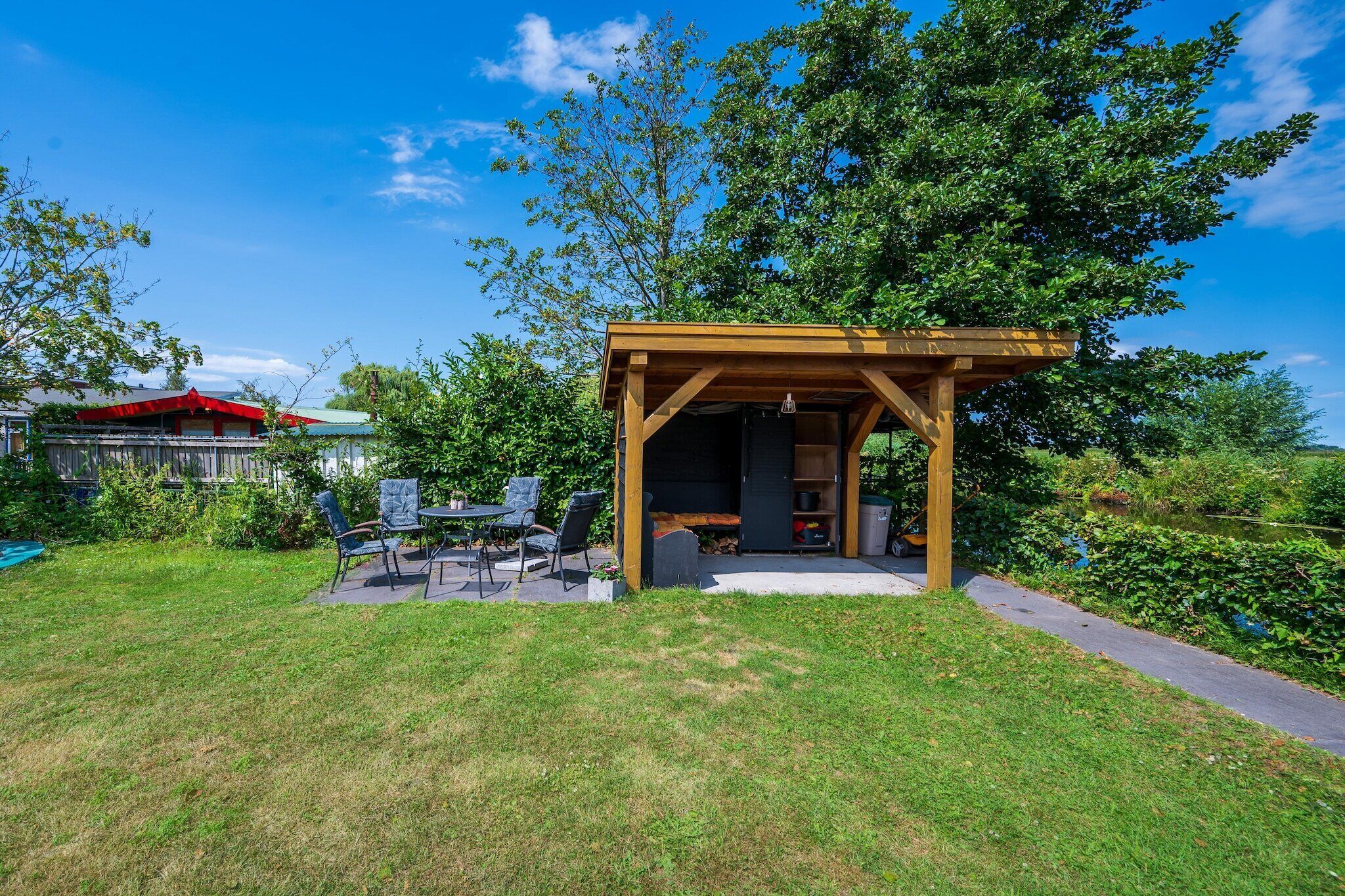 Een houten pergola met een tafel en stoelen, een tuinhuis met een deur, en een gazon.