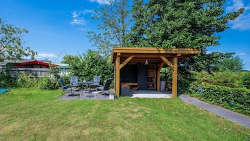 Een houten pergola met een tafel en stoelen, een tuinhuis met een deur, en een gazon.