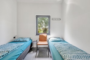 A small room with two single beds, a chair, a window, and a wall-mounted light switch.