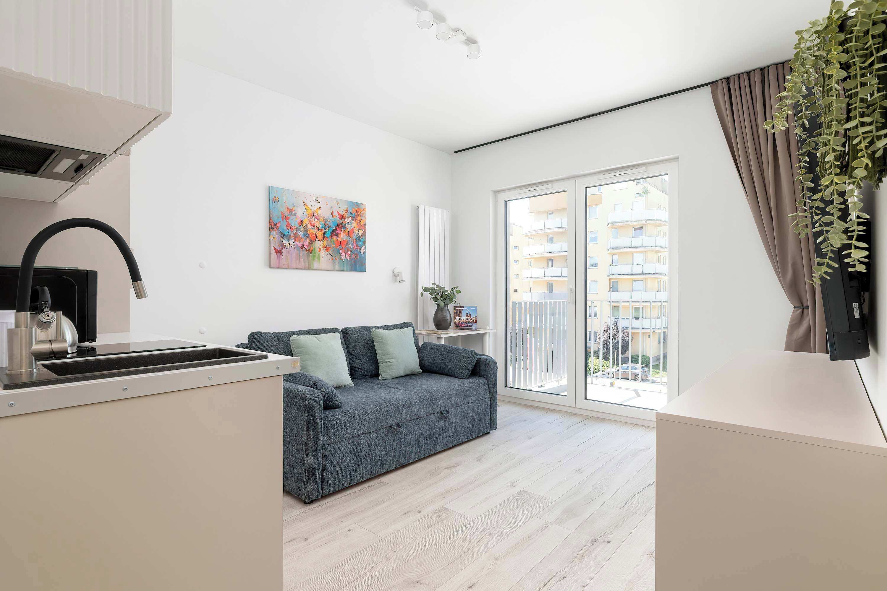 Un appartement moderne doté d’une kitchenette, d’un canapé gris, d’un tableau coloré et offrant une vue sur un immeuble d’habitation.