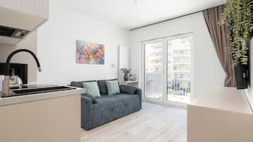 A modern apartment with a kitchenette, a grey sofa, a colorful painting, and a view of a residential building.