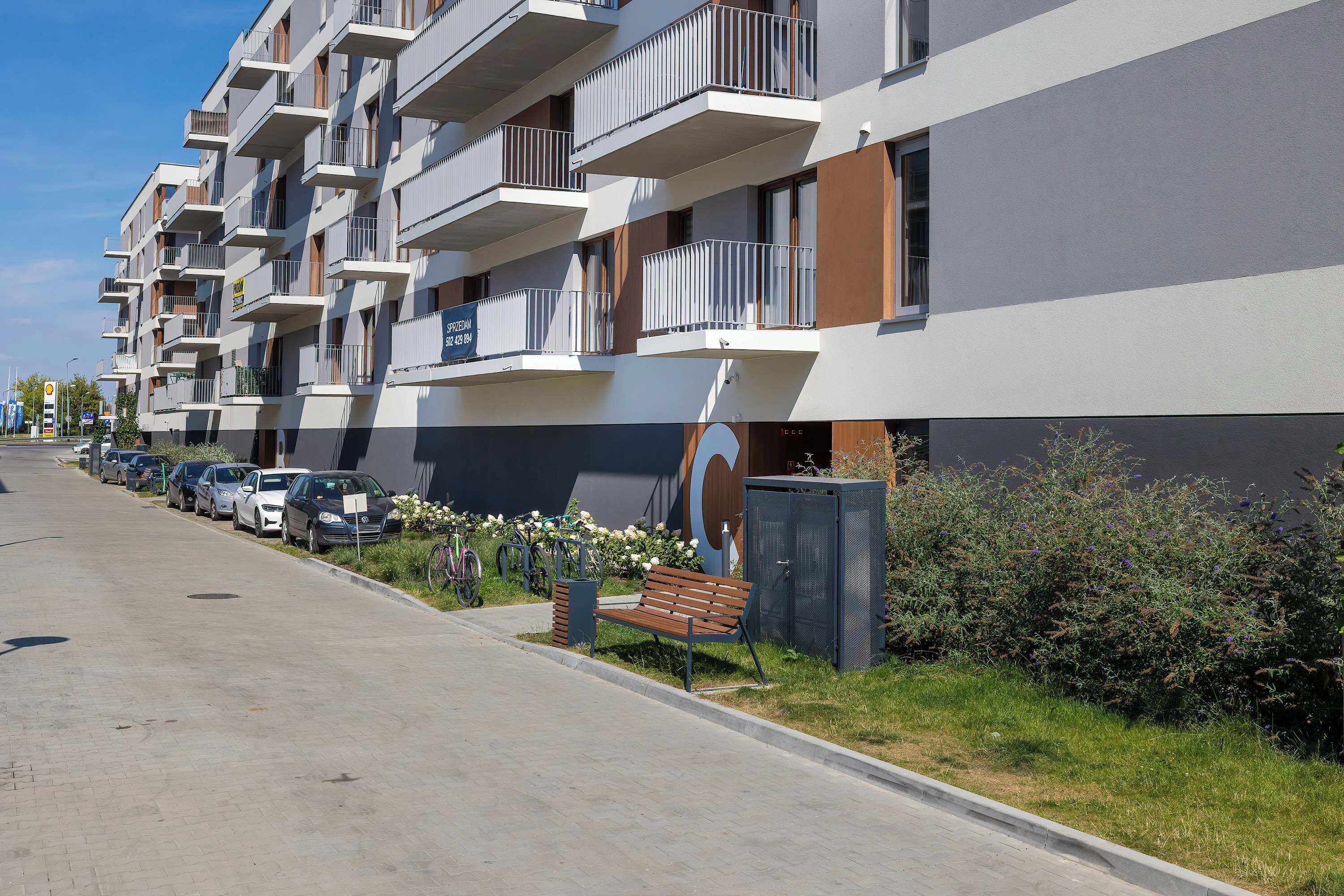 Um edifício de apartamentos moderno com várias varandas, área de estacionamento com carros e um pequeno jardim com um banco.