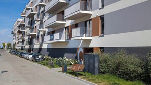 Un immeuble d’appartements moderne doté de nombreux balcons, d’un espace de stationnement avec des voitures et d’un petit jardin agrémenté d’un banc.