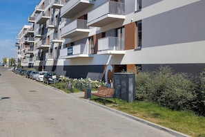 Um edifício de apartamentos moderno com várias varandas, área de estacionamento com carros e um pequeno jardim com um banco.
