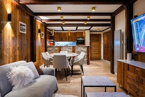 A modern kitchen with wooden accents, a dining area with a round table and chairs, and a living space with a sofa and a television.