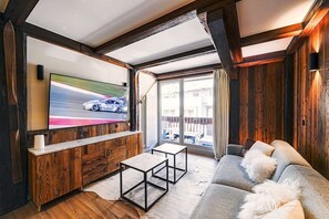 Een moderne woonkamer met een grote flatscreen televisie bevestigd aan een houten wand, een grijze bank met witte kussens en een houten salontafel.