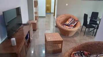 A living room with a flat-screen TV, a wooden cabinet, a wicker chair with a cushion, and a small table.