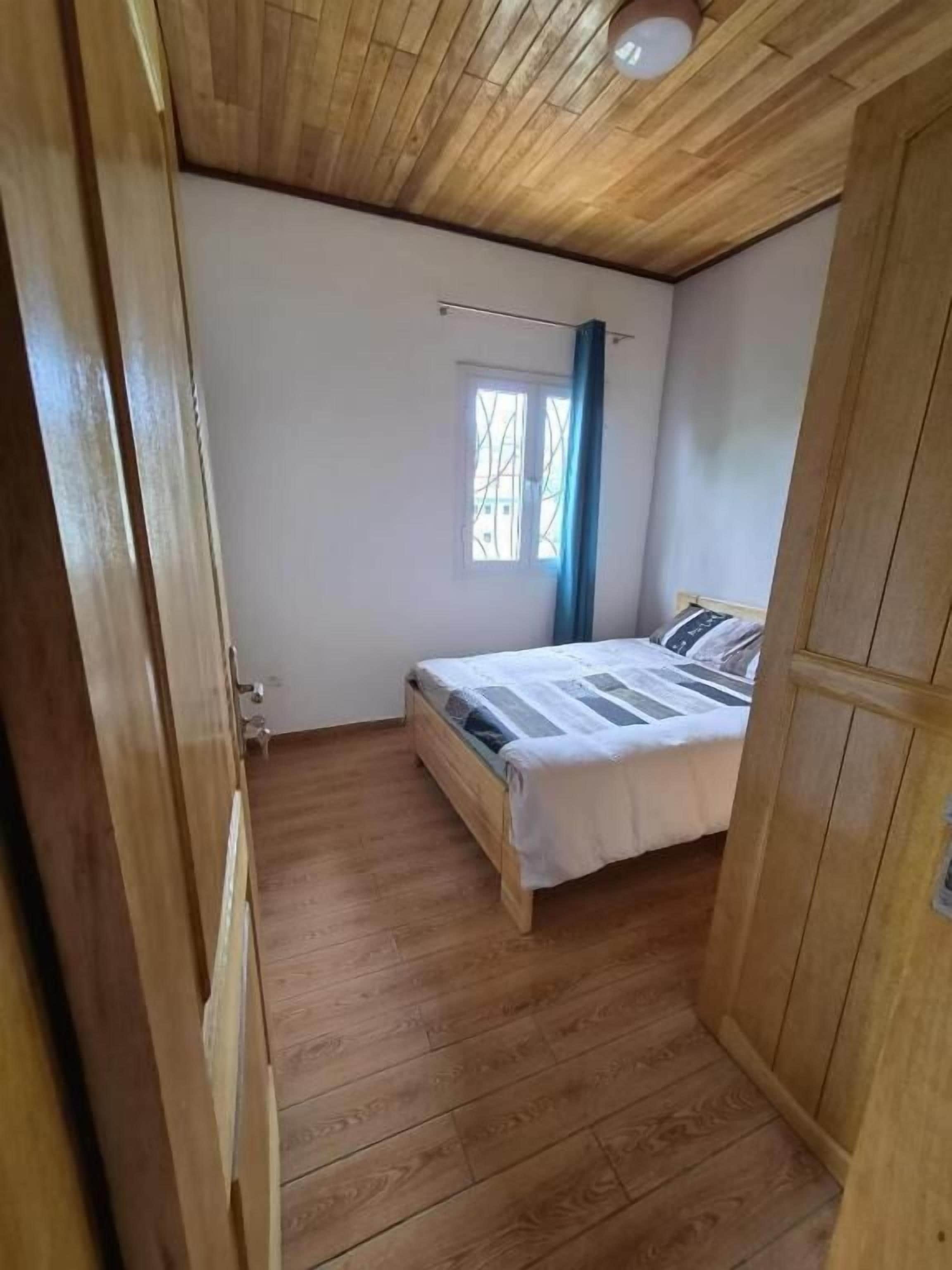 A bedroom with a wooden ceiling, a bed with a white bedspread, a window with blue curtains, and a wooden wardrobe.