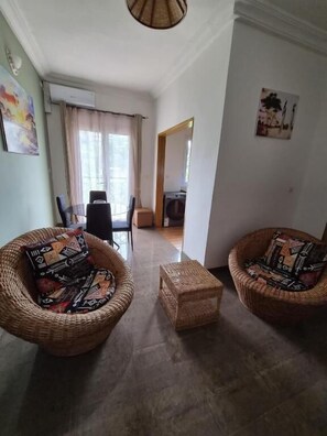 Un salon avec deux fauteuils en osier, une petite table et une porte-fenêtre donnant sur une autre pièce.