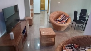 A living room with a flat-screen TV, a wooden cabinet, a wicker chair with a cushion, and a small table. - Charming 2-bedroom Apartment in Yaoundé - Comfortable Living (Yaoundé)