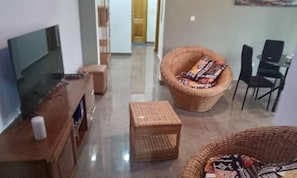 A living room with a flat-screen TV, a wooden cabinet, a wicker chair with a cushion, and a small table. - Charming 2-bedroom Apartment in Yaoundé - Comfortable Living (Yaoundé)