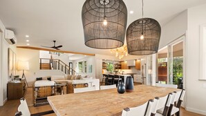 Een ruime woonkamer met een grote houten eettafel, moderne hanglampen, en uitzicht op de keuken en buitenruimte.