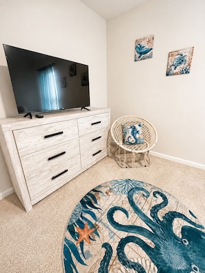 A room with a television on a wooden cabinet, a woven chair with a cushion, and a round rug with a sea creature design. - Enjoy our newly built 5-bedroom, 4.5-bath home just 3 miles from Holden Beach. (Supply)