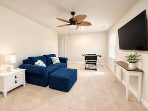 Ein Wohnzimmer mit einem blauen Sofa, einem Hocker und einem Kicker. Es gibt einen Deckenventilator, einen an der Wand montierten Fernseher und einen weißen Nachttisch mit einer Lampe.