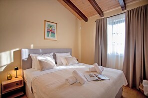 Een net opgemaakt bed met kussens en een opgerolde handdoek, een nachtkastje met een lamp, een ingelijst beeld aan de muur, en een raam met gordijnen.