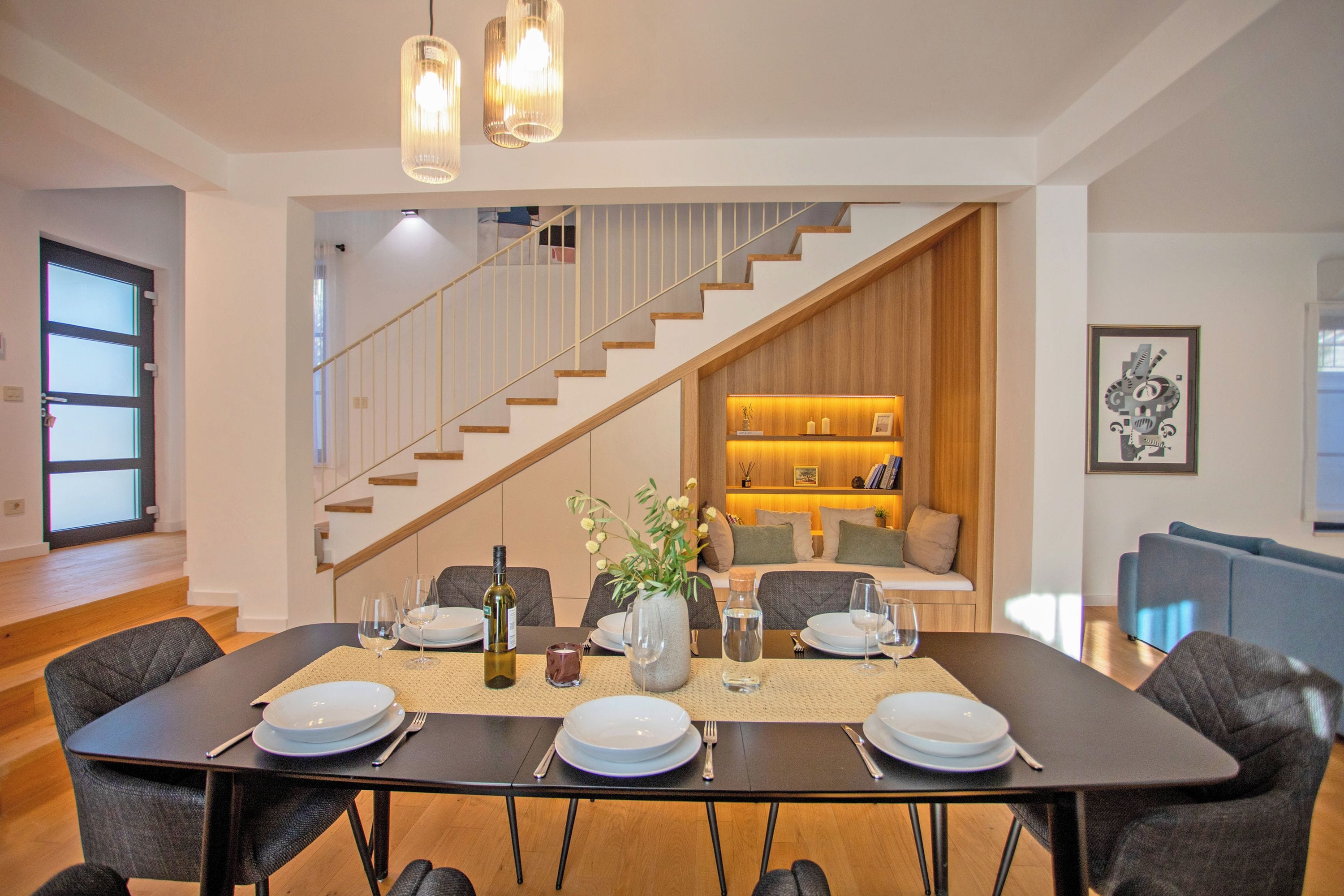 Un comedor con una mesa dispuesta para cuatro, una escalera y una sala de estar con sofá y estanterías.