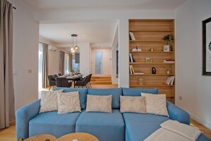 Ein Wohnzimmer mit einem blauen Sofa, einem Holzregal und einem Essbereich inklusive Tisch und Stühlen.