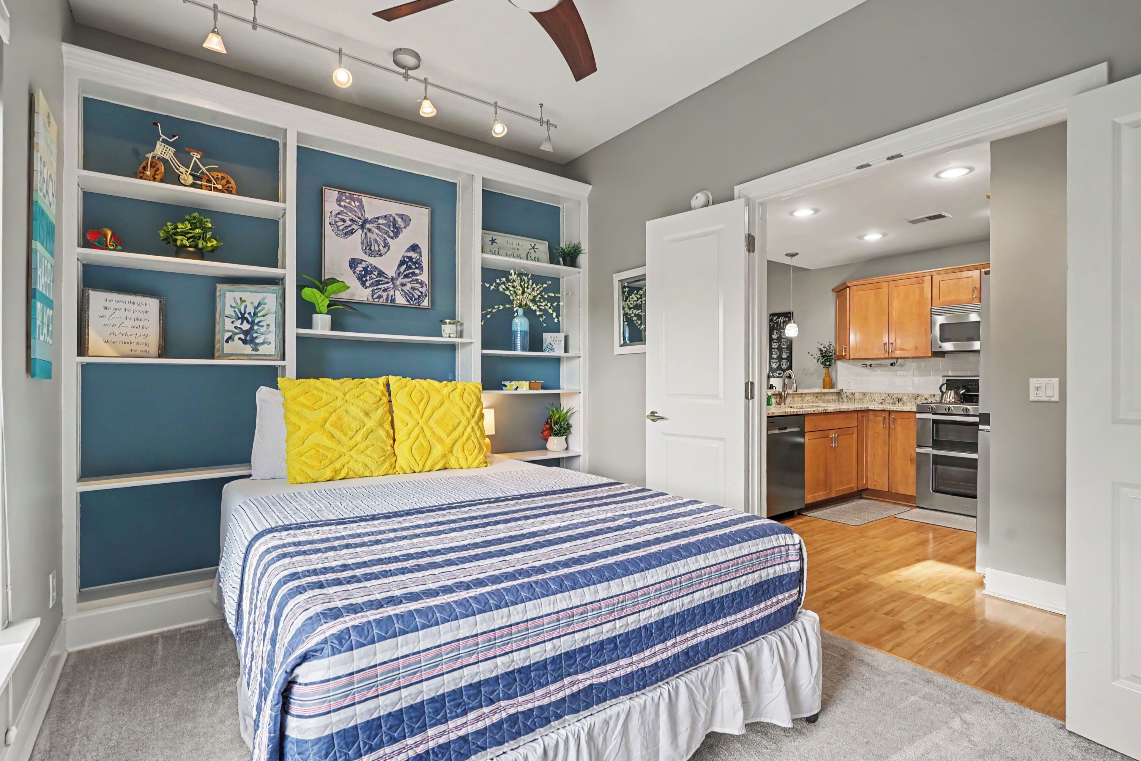 A bedroom with a bed, a nightstand, a dresser, and a wall with shelves containing various items.