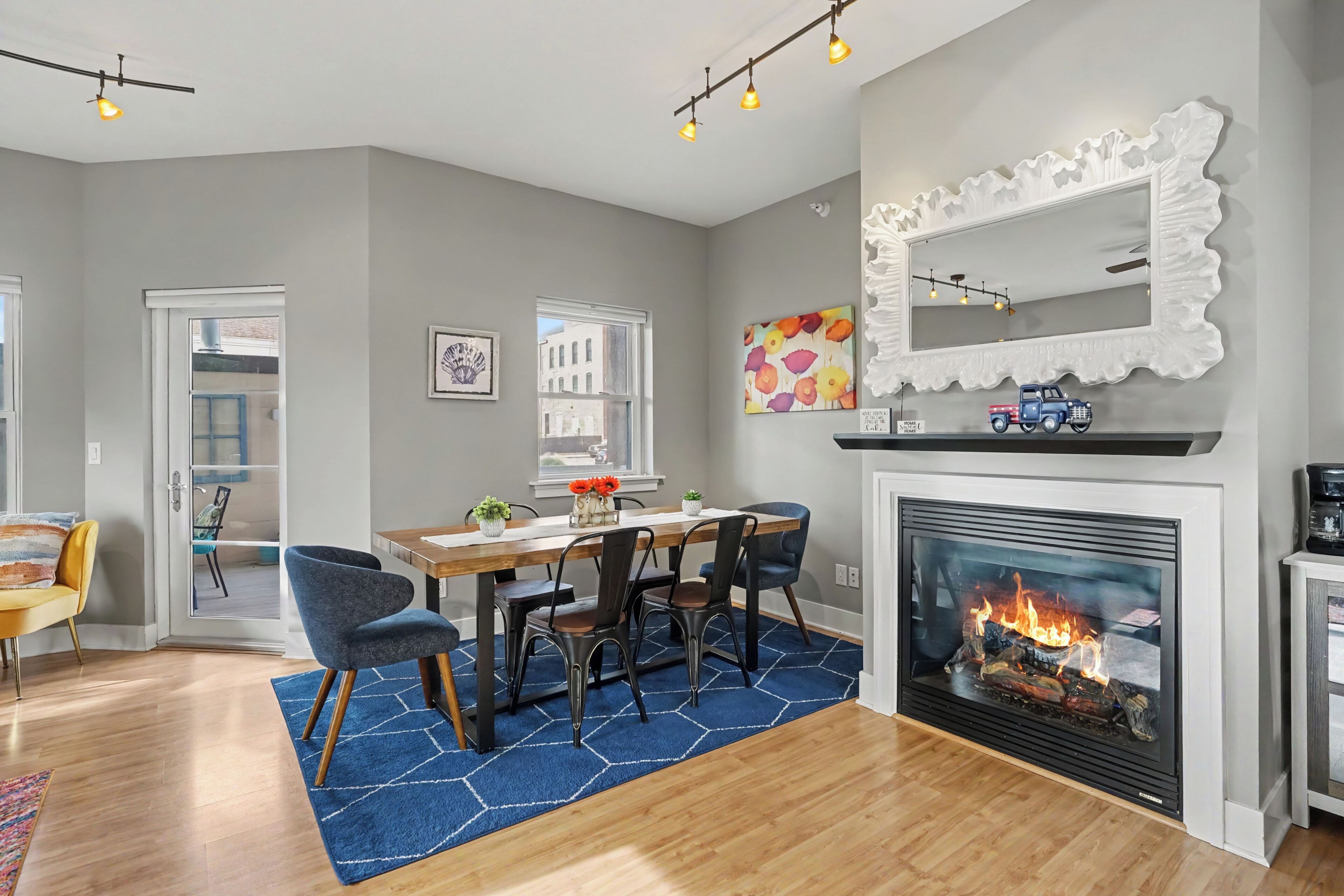 A dining area with a wooden table, chairs, and a blue rug. There is a fireplace with a visible fire and a decorative mirror above it.