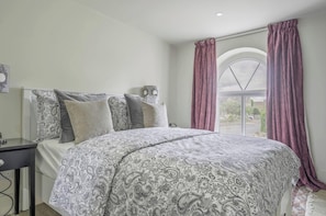 A bedroom with a bed, a nightstand, a window with curtains, and a view of the outside. - Old Chapel in the heart of South Cerney, charming house with a beautiful garden (South Cerney)