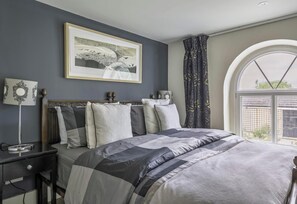 A bedroom with a large bed, a bedside table, a lamp, a framed picture on the wall, and a window with curtains. - Old Chapel in the heart of South Cerney, charming house with a beautiful garden (South Cerney)