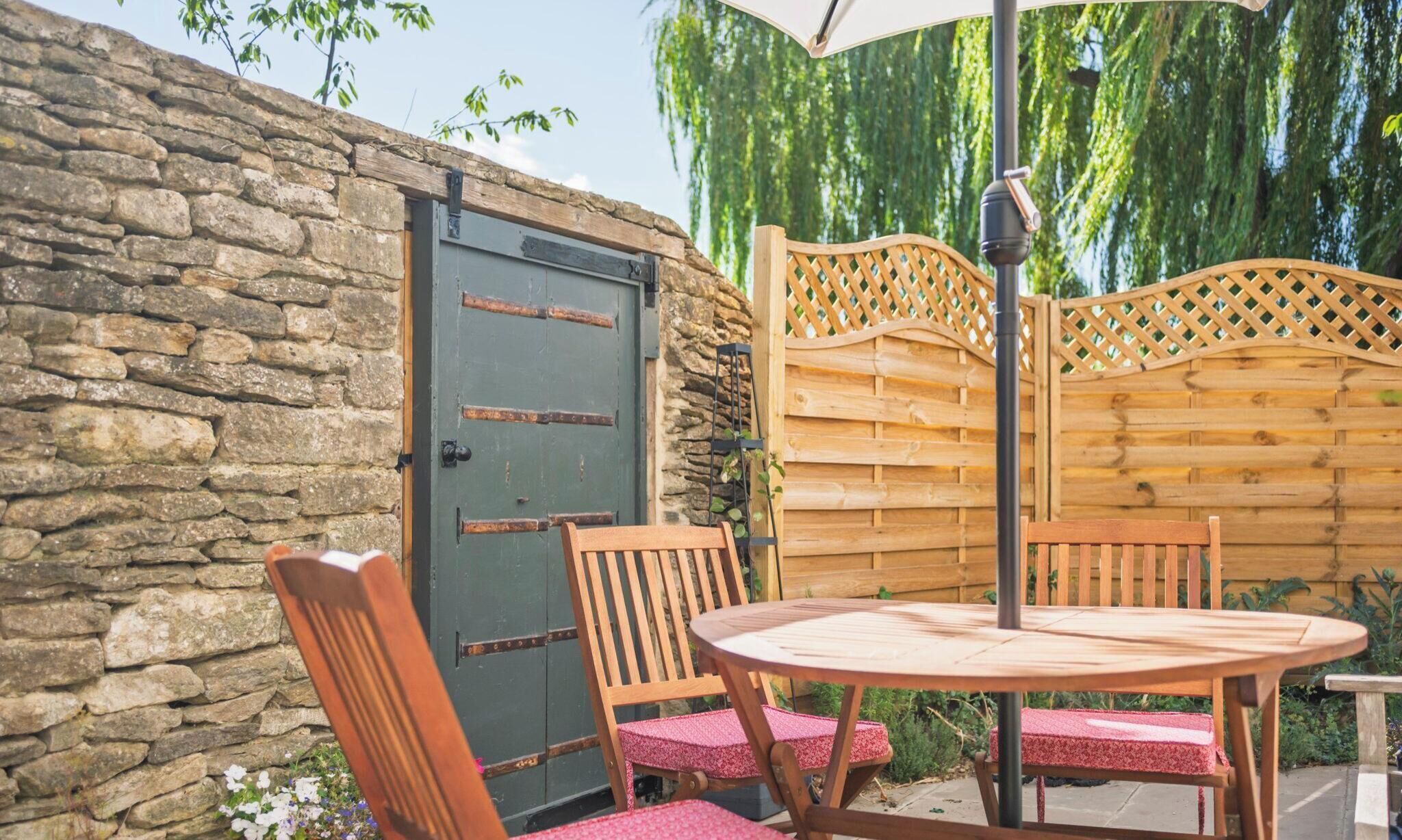 Een stenen muur, een houten poort, een houten omheining en een ronde tafel met stoelen.