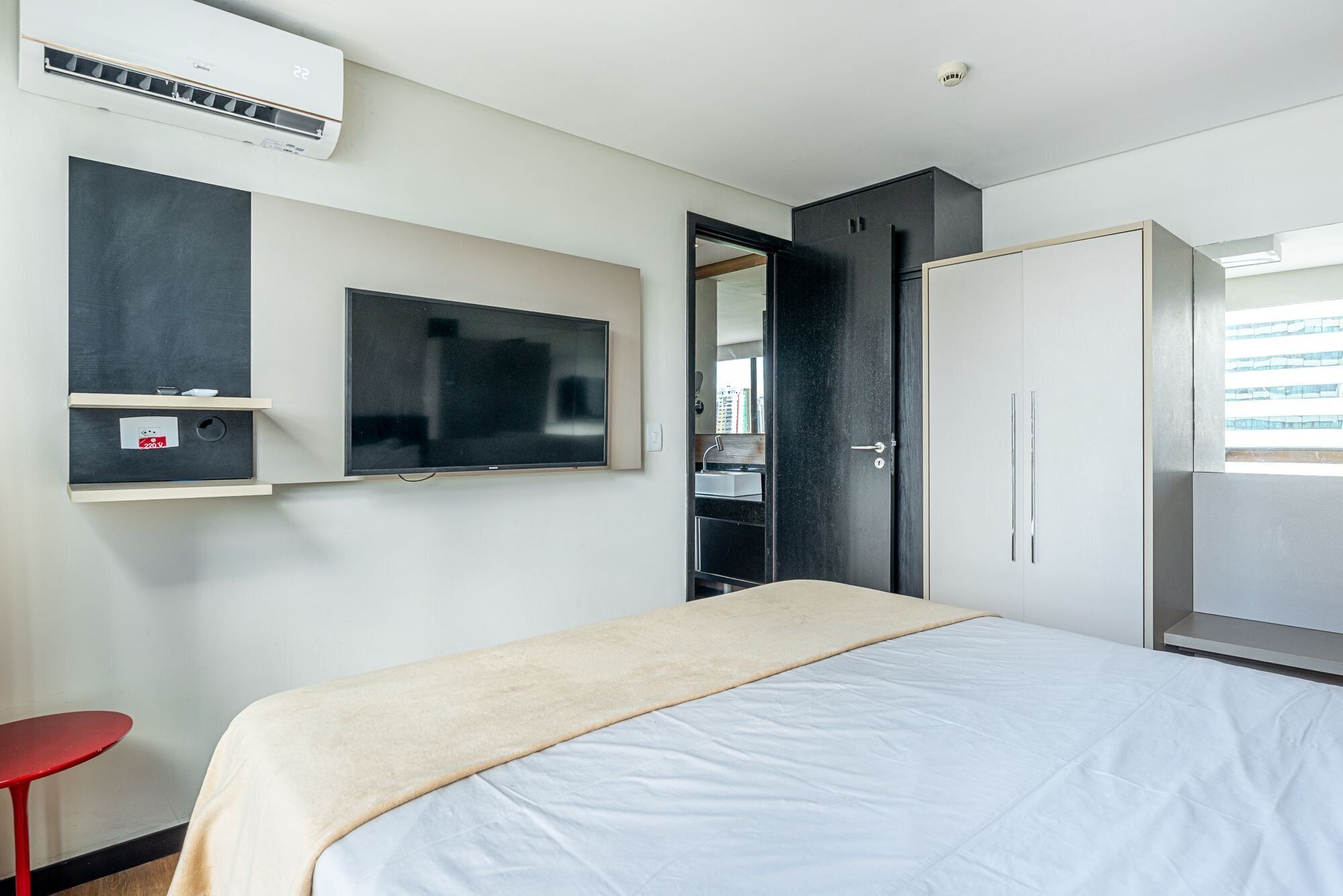 A hotel room with a bed, a TV mounted on the wall, a wardrobe, and a red chair.