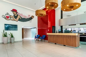 A modern reception area with a wooden reception desk, blue chairs, and a mural with a person watering a plant. - Aparament em Boa Viagem-Prox a Praia-167 (Recife)