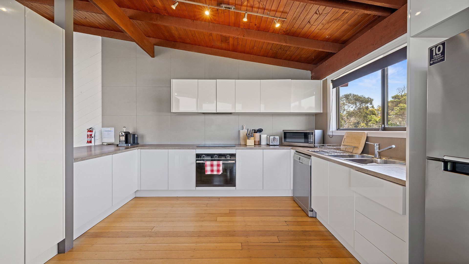 A modern kitchen with white cabinets, stainless steel appliances, a wooden ceiling, and a window providing a view of trees.