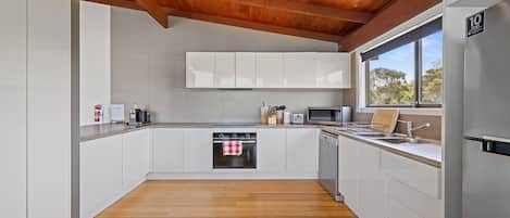 A modern kitchen with white cabinets, stainless steel appliances, a wooden ceiling, and a window providing a view of trees.