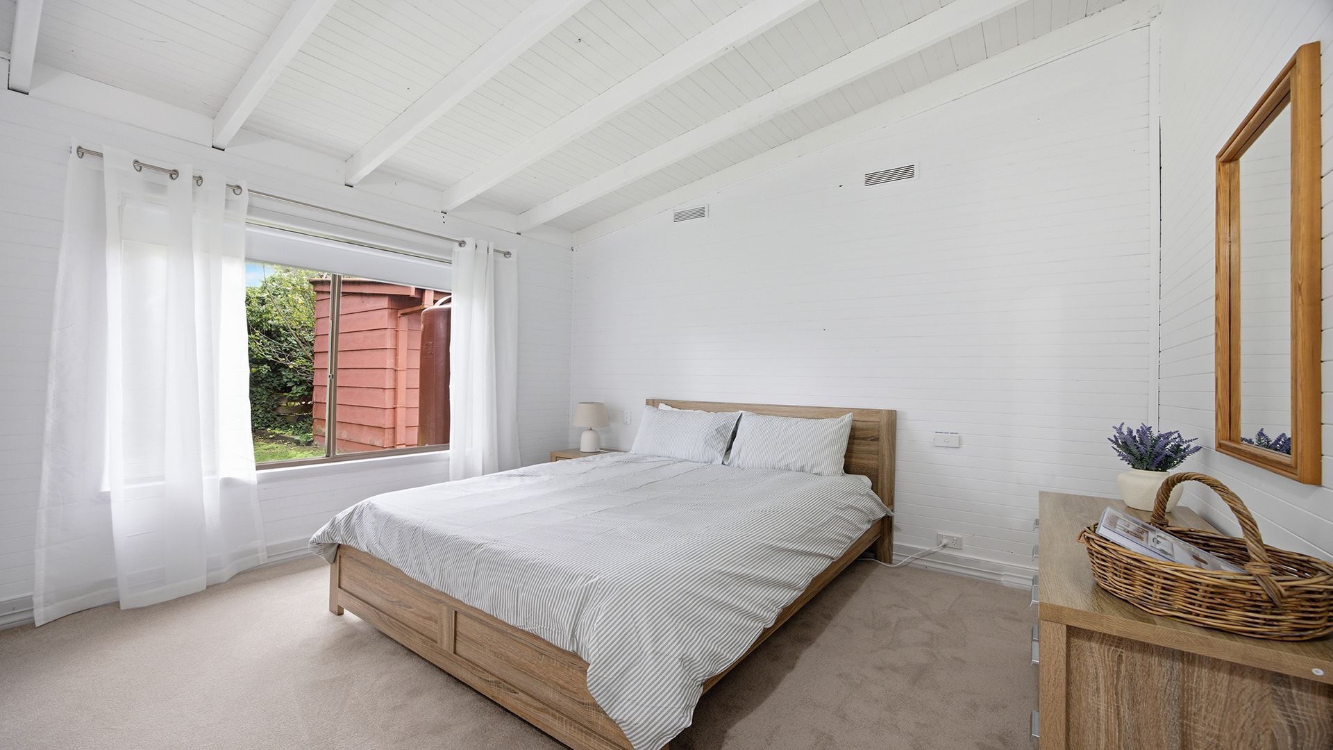 Ein Schlafzimmer mit einem hölzernen Bett, einem Nachttisch, einem Spiegel und einem Fenster mit transparenten Vorhängen.
