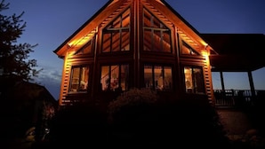 Une cabane en rondins avec un toit triangulaire et de grandes fenêtres, entourée d’une terrasse et d’un espace abrité.