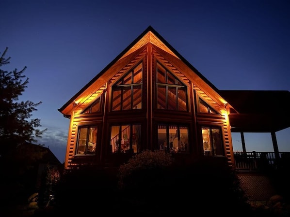 A log cabin with a triangular roof and large windows, surrounded by a deck and a covered area.