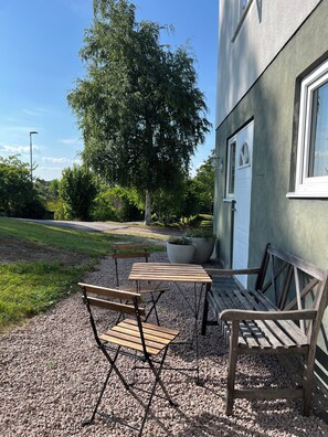 An outdoor seating area with wooden tables and benches, gravel ground, and a building with a window and door. - Sjöbacken – Lovely apartment in the heart of Braås (Braås)