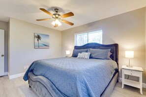 Ein Schlafzimmer mit Bett, Deckenventilator, Nachttisch, Lampe, Fenster und Wandbild.